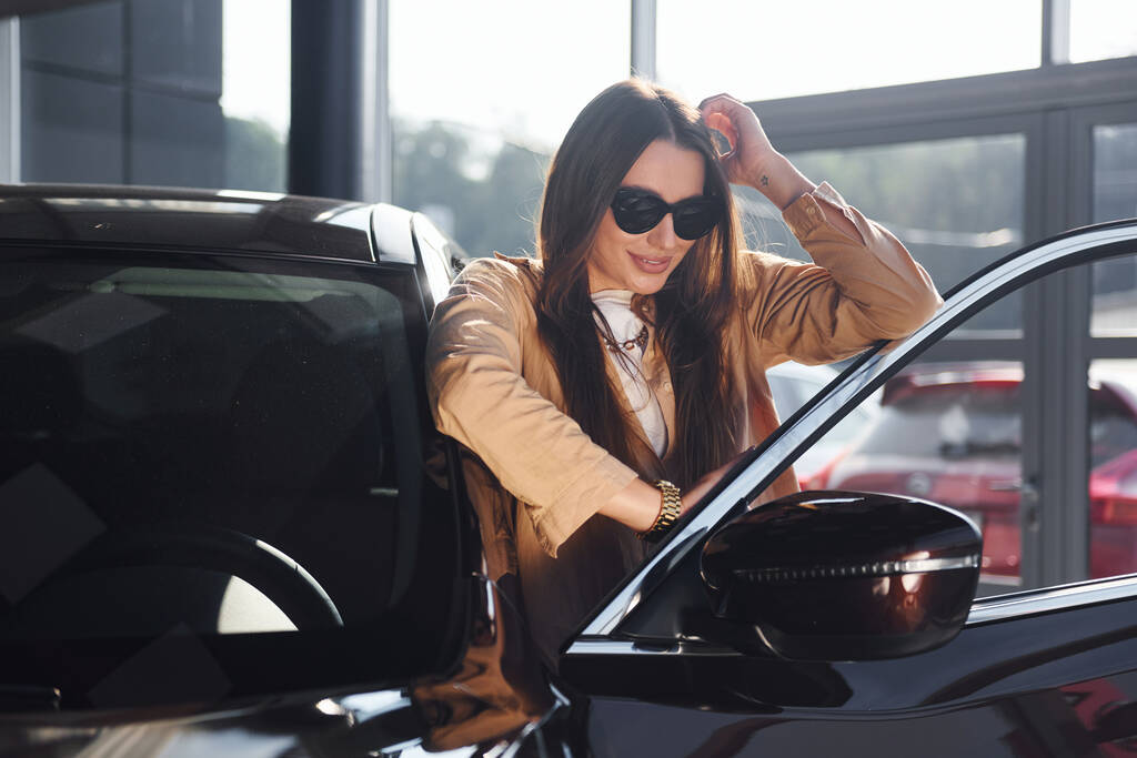 In der Nähe eines geöffneten Fensters stehend. Modisch schöne junge Frau und ihr modernes Automobil. - Foto, Bild