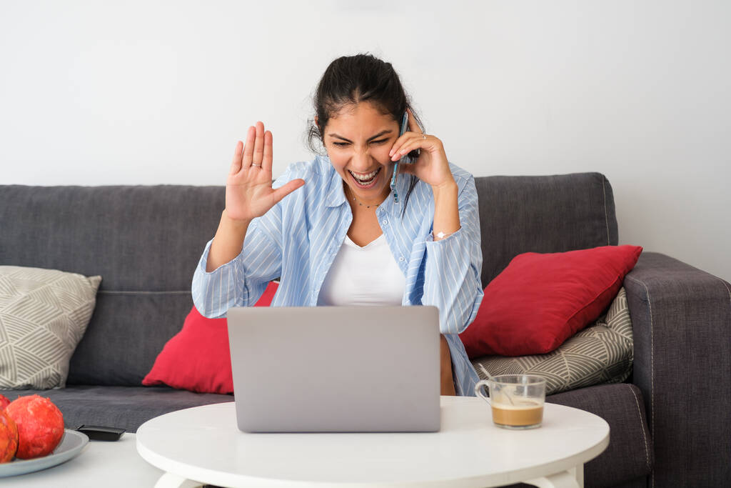 Young excited latin casual woman on mobile phone call using laptop at home. Expressive girl gesturing. - Photo, Image