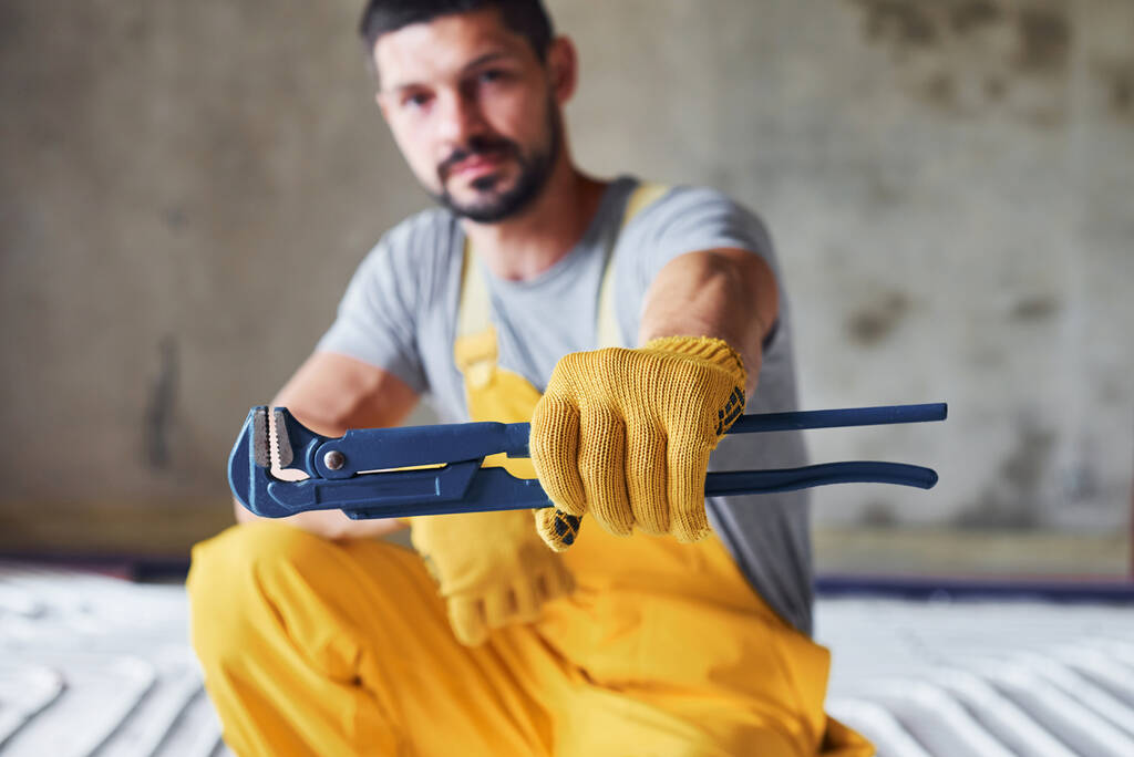 Mantiene la llave en la mano. Trabajador en uniforme de color amarillo instalando sistema de calefacción por suelo radiante. - Foto, Imagen