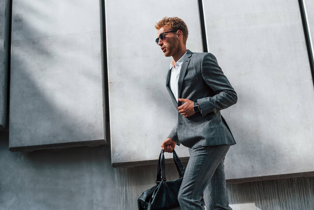 In sunglasses. Young successful businessman in grey formal wear is outdoors in the city. - Photo, Image