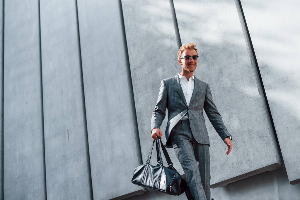 В солнечных очках. Молодой успешный бизнесмен в серой формальной одежде на открытом воздухе в городе. - Фото, изображение