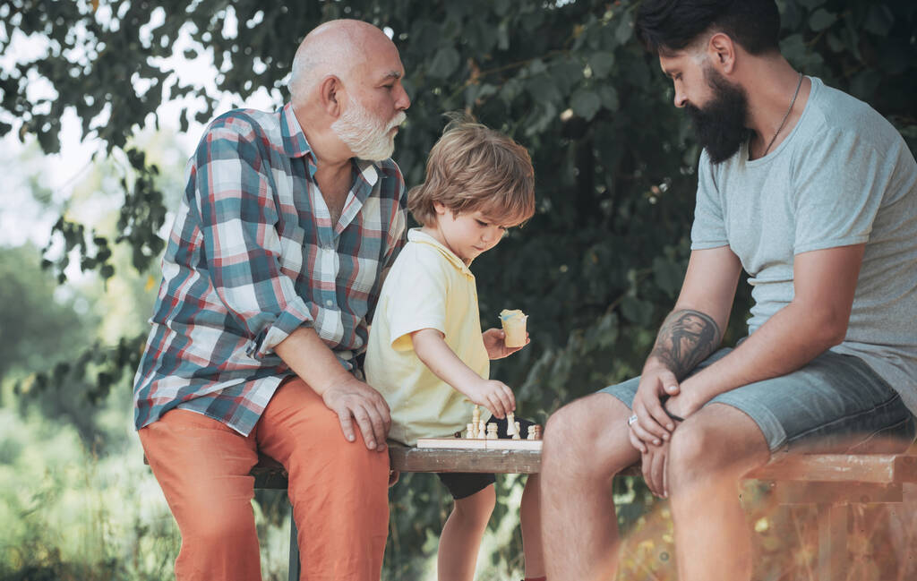 Beau grand-père et petit-fils jouent aux échecs tout en passant du temps ensemble en plein air. Garçon avec père et grand-père. Père et fils dans le parc
. - Photo, image