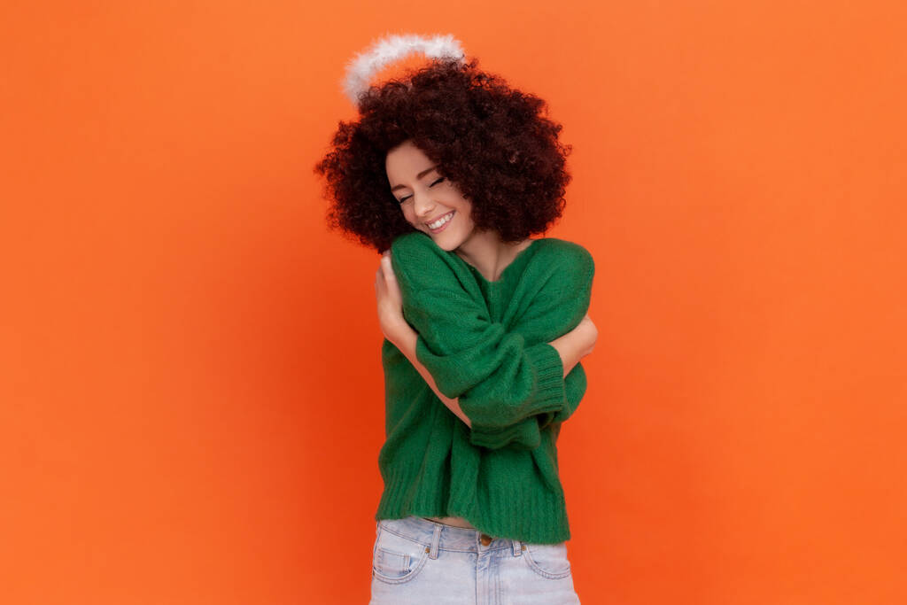 Boldog elégedett nő afro frizurával visel zöld alkalmi stílusú pulóver és nimb feje fölött ölelés magát, kifejező szeretet, ölelést. Beltéri stúdió lövés elszigetelt narancs háttér. - Fotó, kép