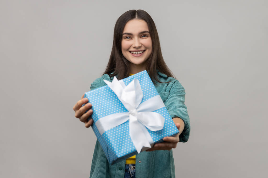 Задоволена молода доросла жінка тримає синю обгорнуту подарункову коробку, даруючи подарунок, дивлячись посміхаючись на камеру, одягнену в повсякденну куртку. Внутрішній студійний знімок ізольований на сірому фоні
. - Фото, зображення