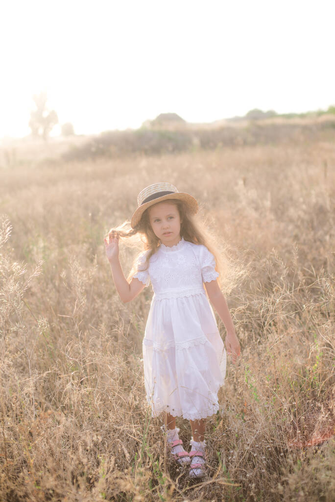 Una graziosa bambina con lunghi capelli biondi ricci in un abito bianco estivo e un cappello di paglia in un campo in campagna in estate al tramonto. Natura ed Ecovita - Foto, immagini