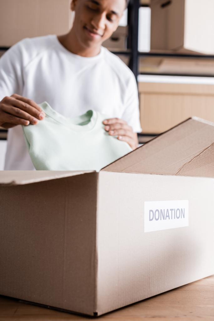 Caja con letras de donación cerca del borroso propietario afroamericano con ropa en la tienda web en línea  - Foto, Imagen