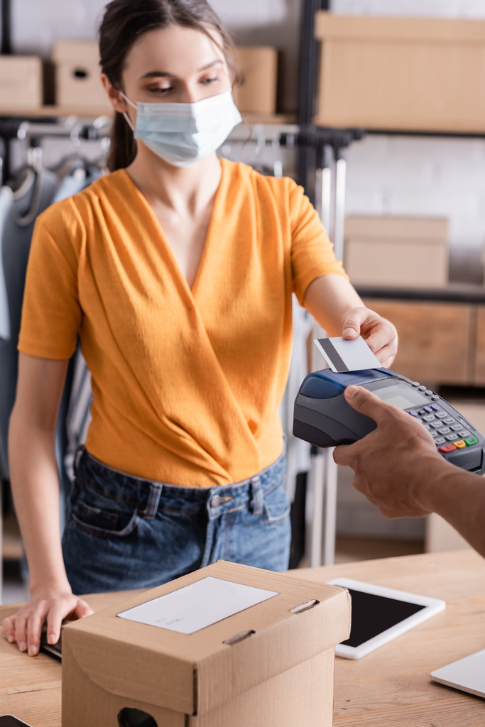 Продавця в медичній масці оплата кредитною карткою біля афроамериканського кур'єра та картонної коробки в інтернет-магазині
  - Фото, зображення