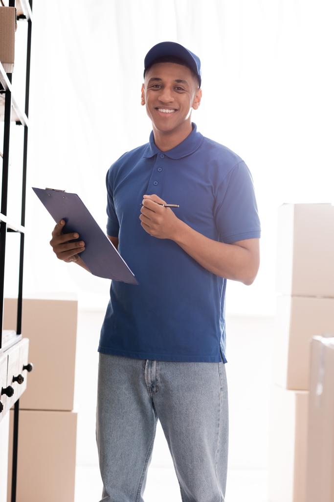 Distribuidor afroamericano positivo sosteniendo portapapeles cerca de cajas de cartón en tienda web en línea  - Foto, imagen