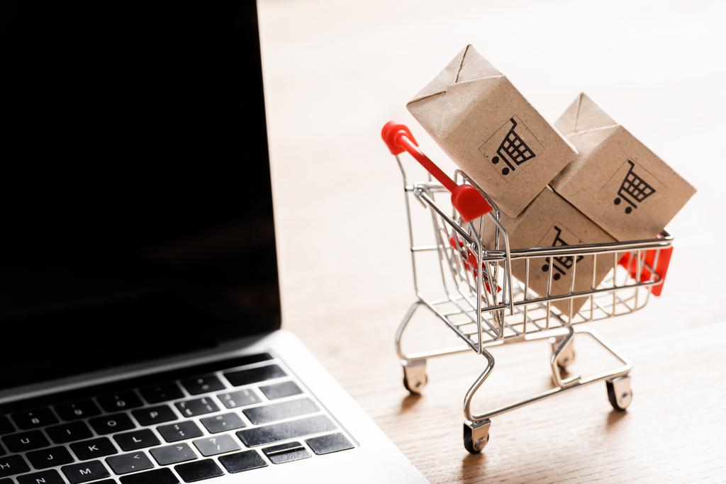 Packages in toy shopping cart near laptop with blank screen on table  - Photo, Image