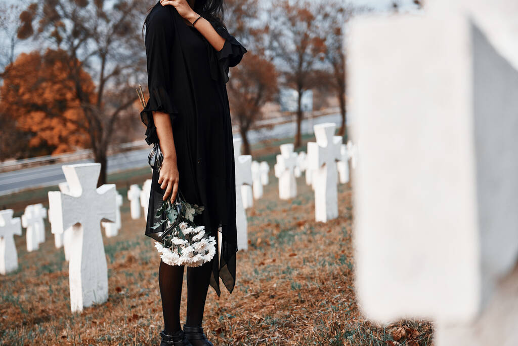 Με λουλούδια στα χέρια. Νεαρή γυναίκα με μαύρα ρούχα επισκέπτεται νεκροταφείο με πολλούς λευκούς σταυρούς. Σύλληψη κηδείας και θανάτου. - Φωτογραφία, εικόνα