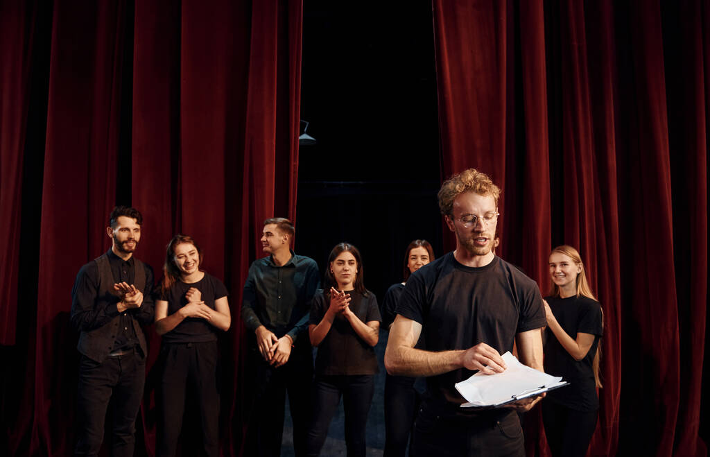 Man met notitieblok oefent zijn rol. Groep acteurs in donker gekleurde kleren op repetitie in het theater. - Foto, afbeelding