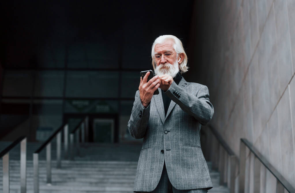 Hombre de negocios senior en ropa formal, con pelo gris y barba está al aire libre probando auriculares inalámbricos con teléfono. - Foto, Imagen