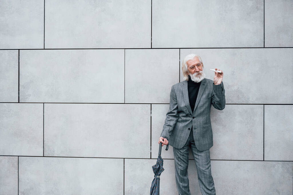 Fumer une cigarette électronique. Homme d'affaires senior en vêtements formels, avec les cheveux gris et la barbe est à l'extérieur. - Photo, image