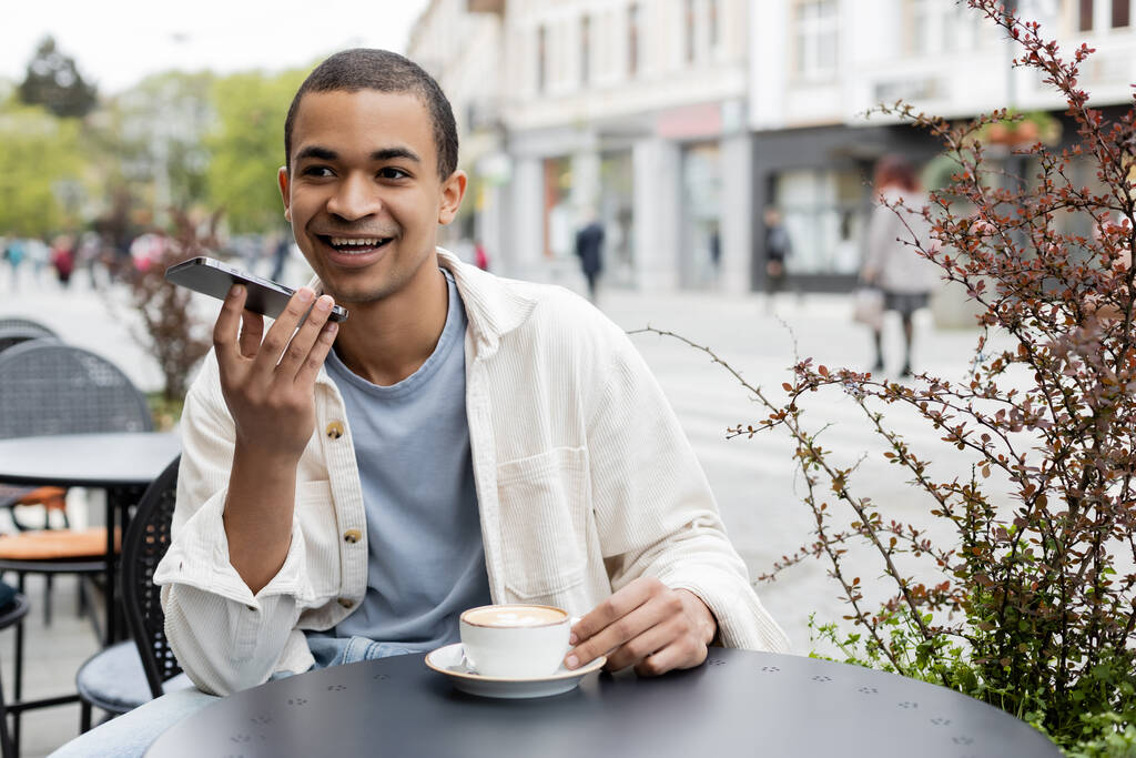 Χαρούμενος Αφροαμερικάνος που ηχογραφεί φωνητικό μήνυμα σε smartphone κοντά σε φλιτζάνι καφέ στο τραπέζι - Φωτογραφία, εικόνα
