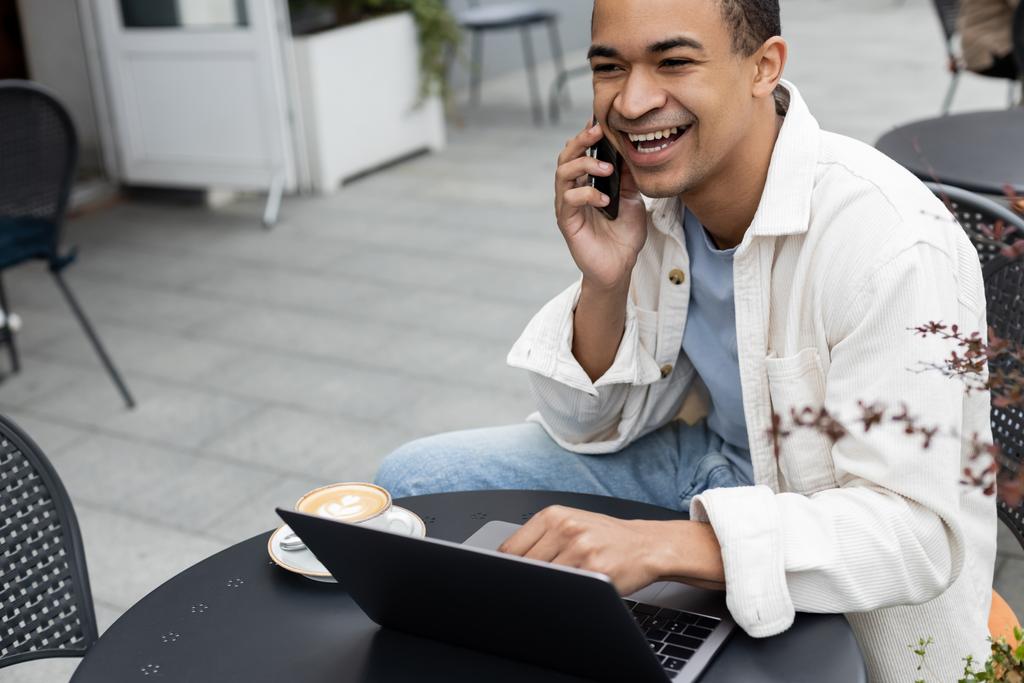 Χαρούμενος Αφρικανός Αμερικανός ελεύθερος επαγγελματίας που μιλάει σε smartphone κοντά στο laptop στη βεράντα του καφέ  - Φωτογραφία, εικόνα