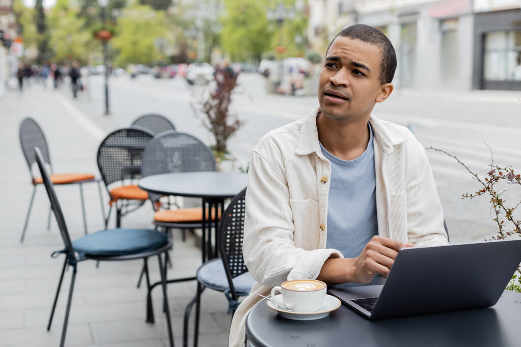 distraído freelancer afroamericano mirando hacia otro lado cerca de la computadora portátil y una taza de capuchino en la terraza de la cafetería  - Foto, imagen