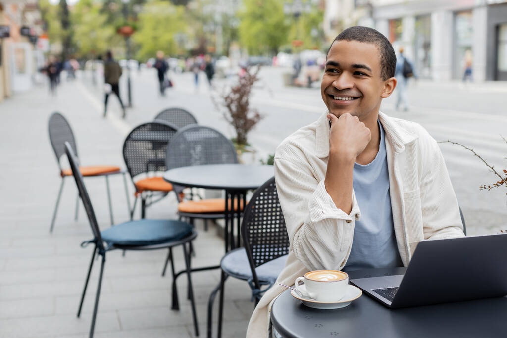 sonriente freelancer afroamericano mirando hacia otro lado cerca de la computadora portátil y una taza de capuchino en la terraza de la cafetería  - Foto, Imagen
