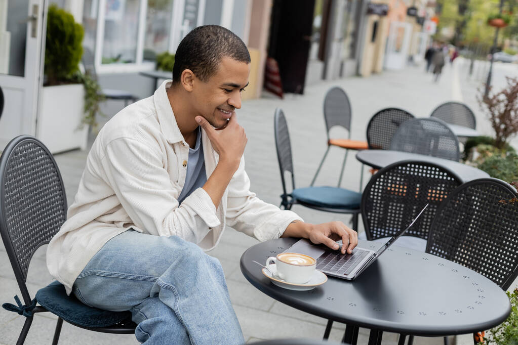 アフリカ系アメリカ人のフリーランスがカフェテラスでカプチーノのカップの近くのノートパソコンでリモートで働いています  - 写真・画像