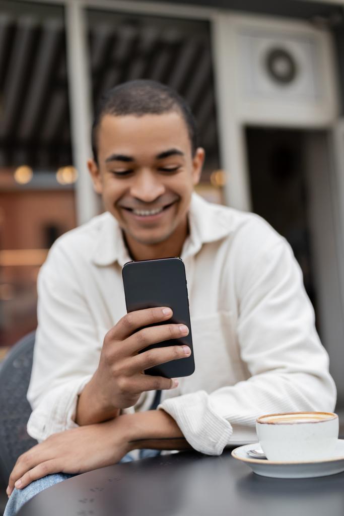 vrolijk Afrikaans amerikaanse man sms 'en op smartphone in de buurt van kopje cappuccino op tafel op cafe terras  - Foto, afbeelding