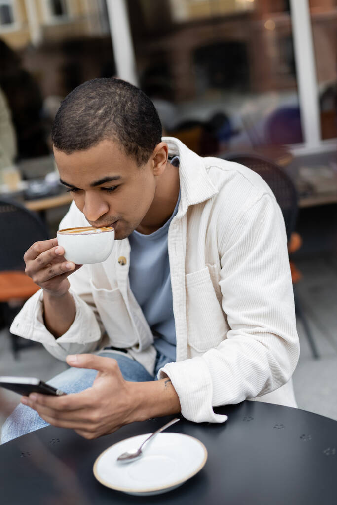 african american man texting on smartphone and drinking cappuccino on cafe terrace  - Photo, Image