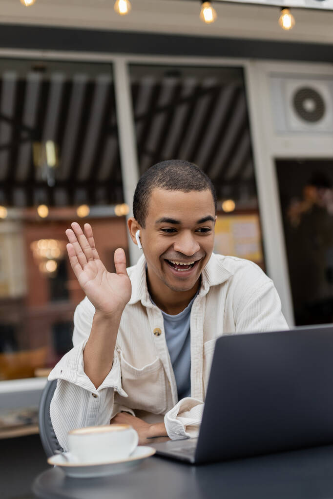 gelukkig Afrikaans amerikaanse man zwaaien hand tijdens video oproep op laptop op zomerterras in cafe - Foto, afbeelding