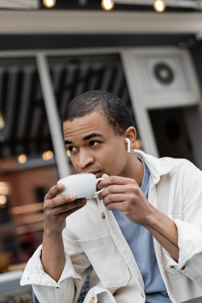 Afrika kökenli Amerikalı kablosuz kulaklıklı adam yaz terasında kahve içiyor. - Fotoğraf, Görsel