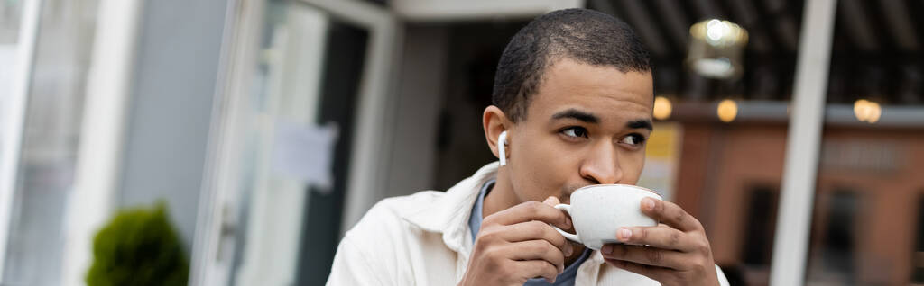 Afrikkalainen amerikkalainen mies langattomissa kuulokkeissa juo kahvia kesäterassilla, banneri - Valokuva, kuva