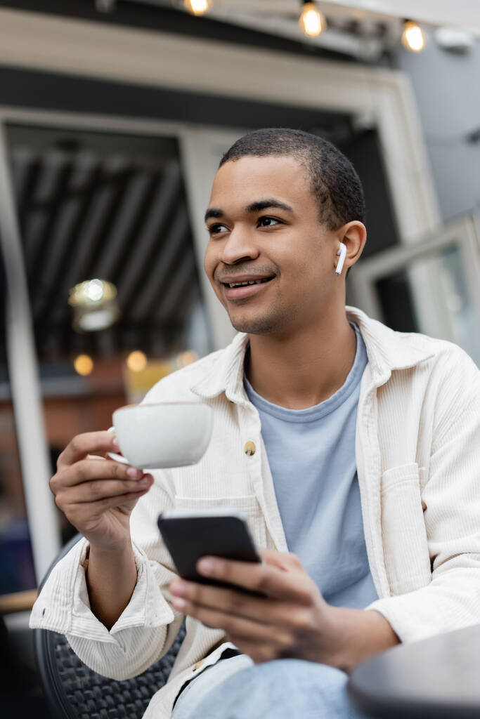 Kablosuz kulaklıklı gülümseyen Afrikalı Amerikalı adam. Elinde bir fincan kahve ve akıllı telefonla yaz terasında.  - Fotoğraf, Görsel