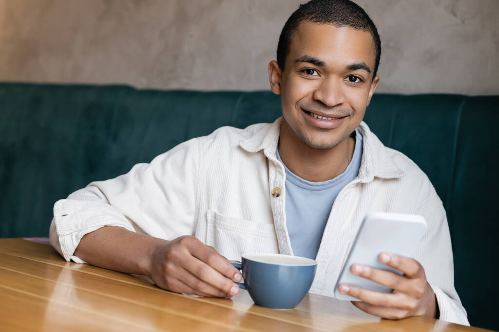 mensajería hombre afroamericano positivo y joven en el teléfono inteligente en la cafetería  - Foto, Imagen