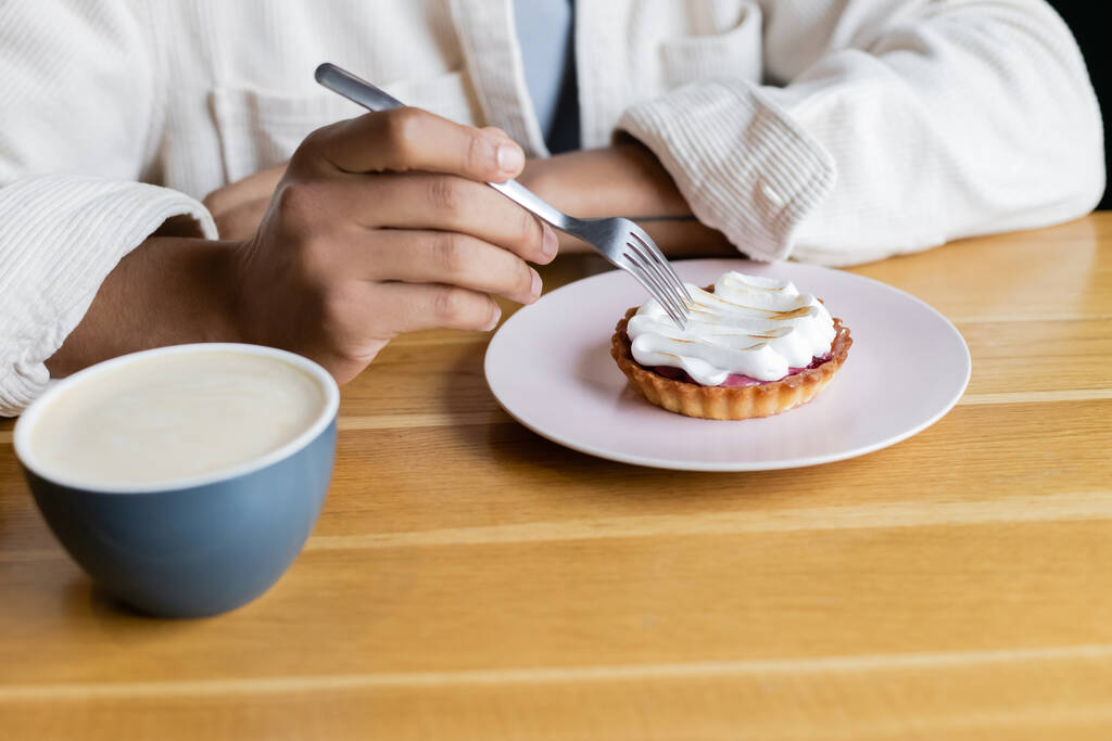 abgeschnittene Ansicht eines afrikanisch-amerikanischen Mannes mit Gabel in der Nähe von Torte und Tasse Cappuccino  - Foto, Bild