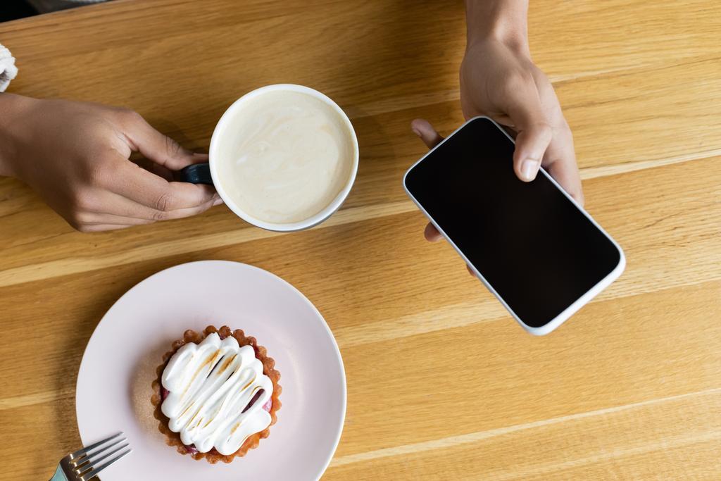 top view of african american man holding smartphone and cup of cappuccino near tasty tart  - Photo, Image