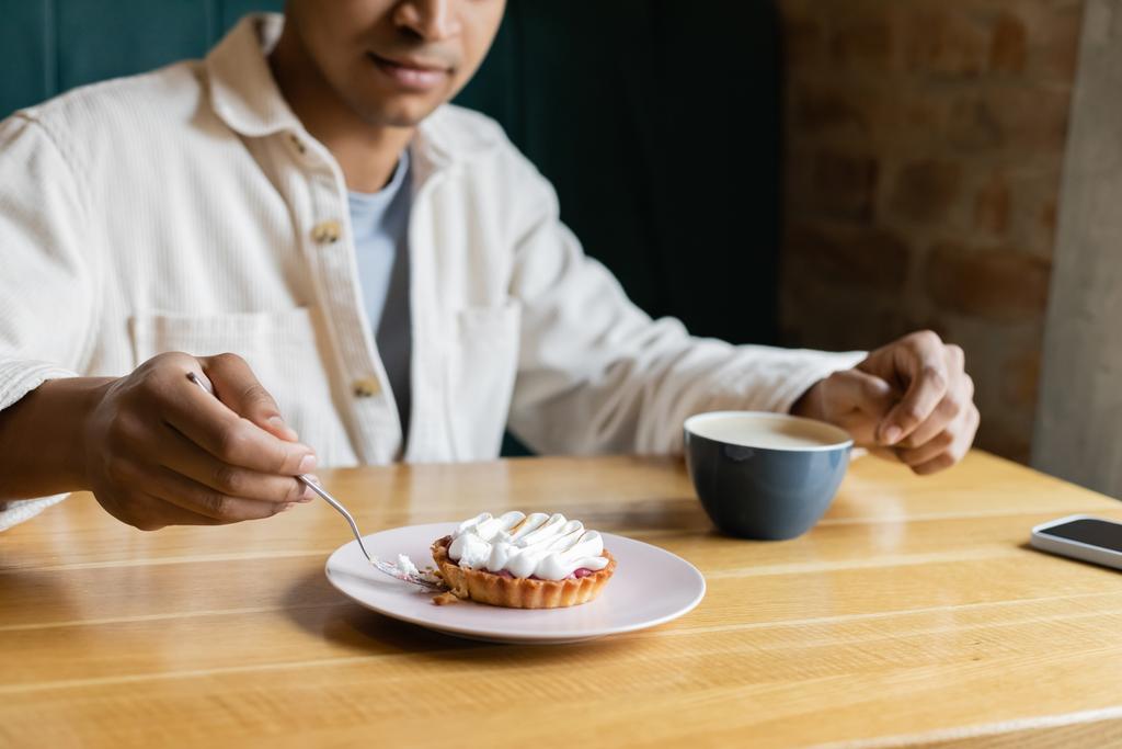 abgeschnittene Ansicht eines jungen afrikanisch-amerikanischen Mannes mit Gabel in der Nähe leckerer Torte auf einem Teller in der Nähe von Tasse und Smartphone - Foto, Bild