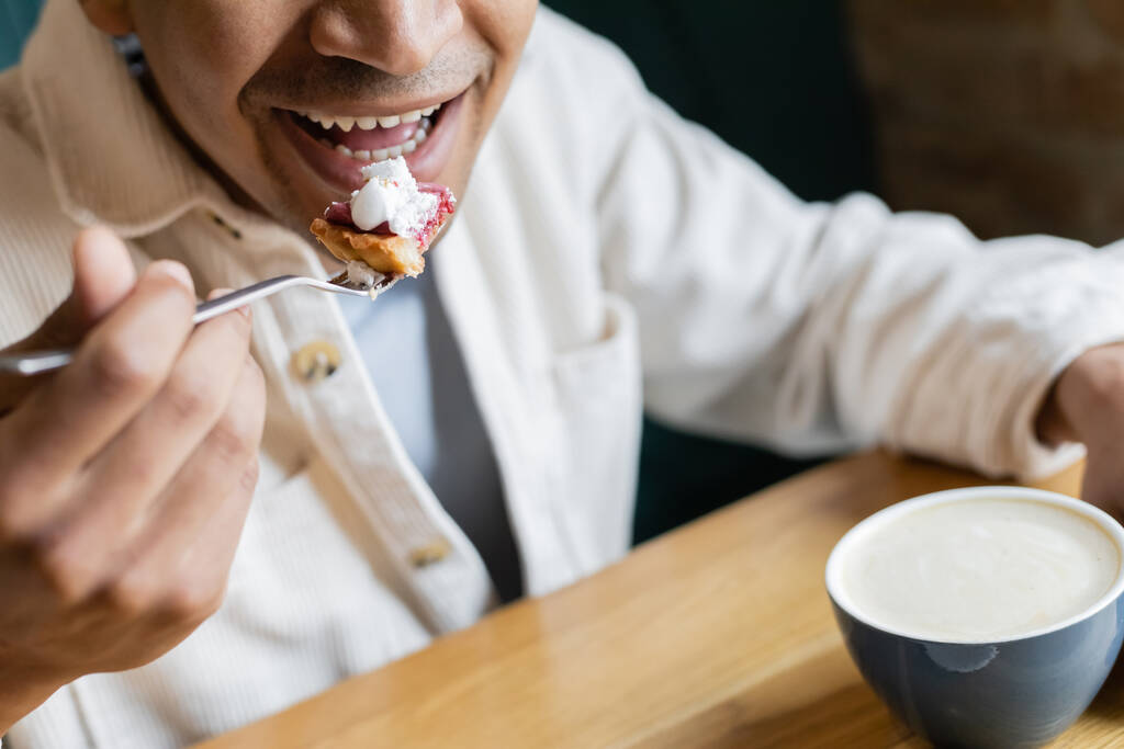コーヒーショップでカプチーノのカップの近くでおいしいケーキを食べる幸せなアフリカ系アメリカ人の男性のクロップドビュー - 写真・画像