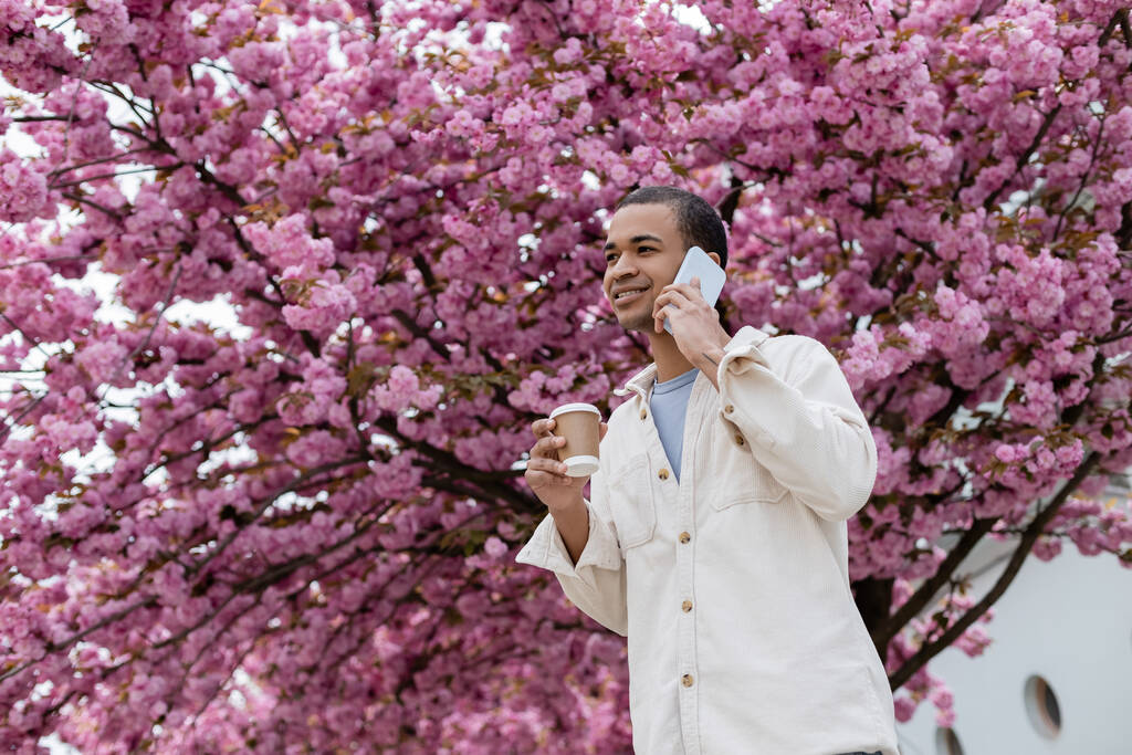 souriant homme afro-américain tenant à emporter boisson et parler sur smartphone près de cerisier rose  - Photo, image