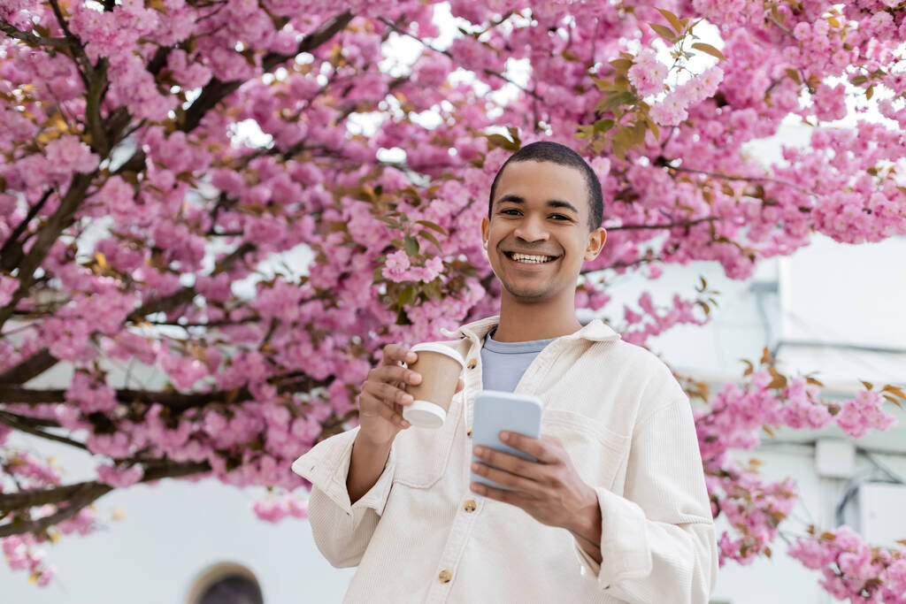 vista a basso angolo di gioioso uomo africano americano che tiene bevanda da asporto e smartphone vicino al ciliegio in fiore  - Foto, immagini