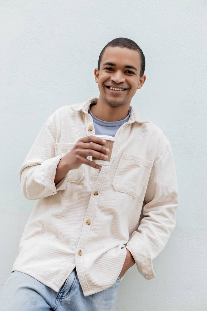 счастливый молодой африканский американец держит бумажный стаканчик возле здания на улице в Европе - Фото, изображение
