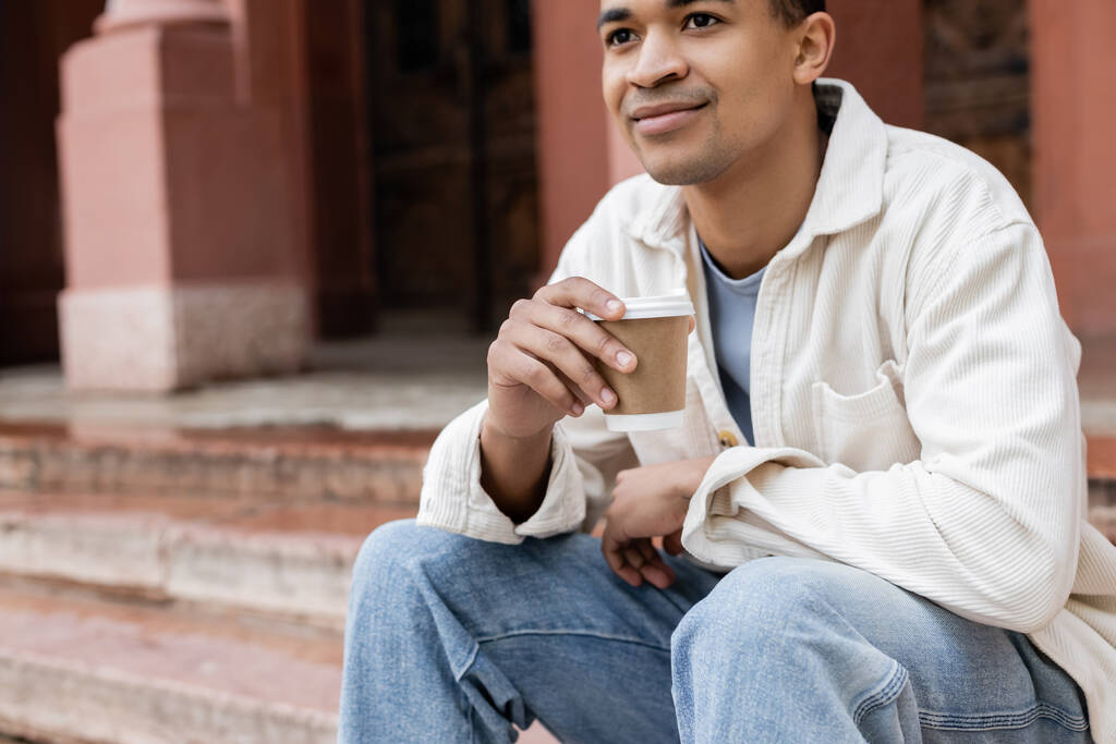 pleased african american man holding takeaway beverage and sitting near building on street in europe - Photo, Image