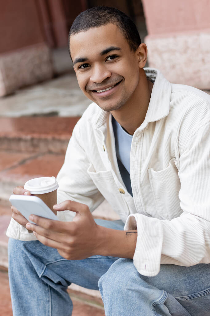 Mutlu Afro-Amerikalı adam elinde içecek ve akıllı telefon tutarken şehirdeki merdivenlerde oturuyor.  - Fotoğraf, Görsel