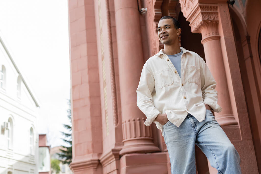 vista basso angolo di piacere afro americano uomo in giacca camicia in piedi con le mani in tasche vicino edificio - Foto, immagini