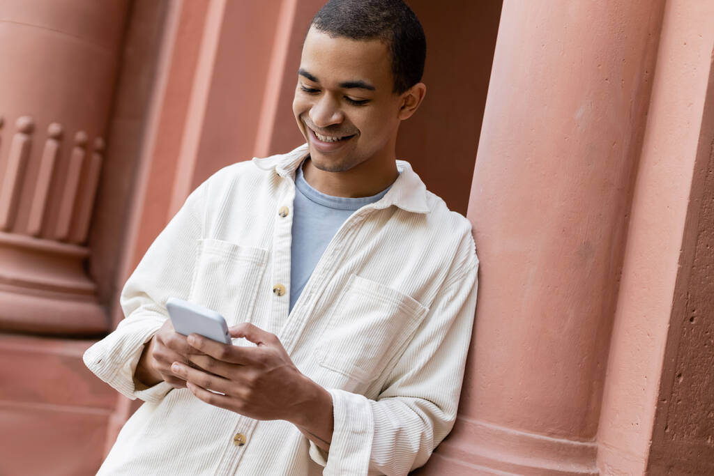 fröhlicher afrikanisch-amerikanischer Mann in Hemdjacke textet auf Smartphone in der Nähe von Gebäude - Foto, Bild