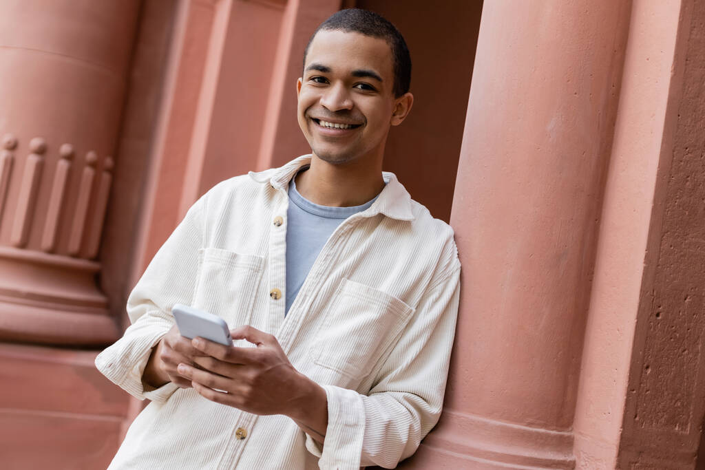 радісний афроамериканський чоловік у сорочці тримає смартфон біля будівлі
 - Фото, зображення