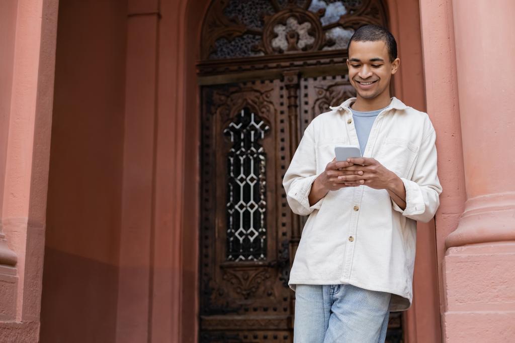 heureux homme afro-américain en veste chemise bavarder sur smartphone près du bâtiment - Photo, image