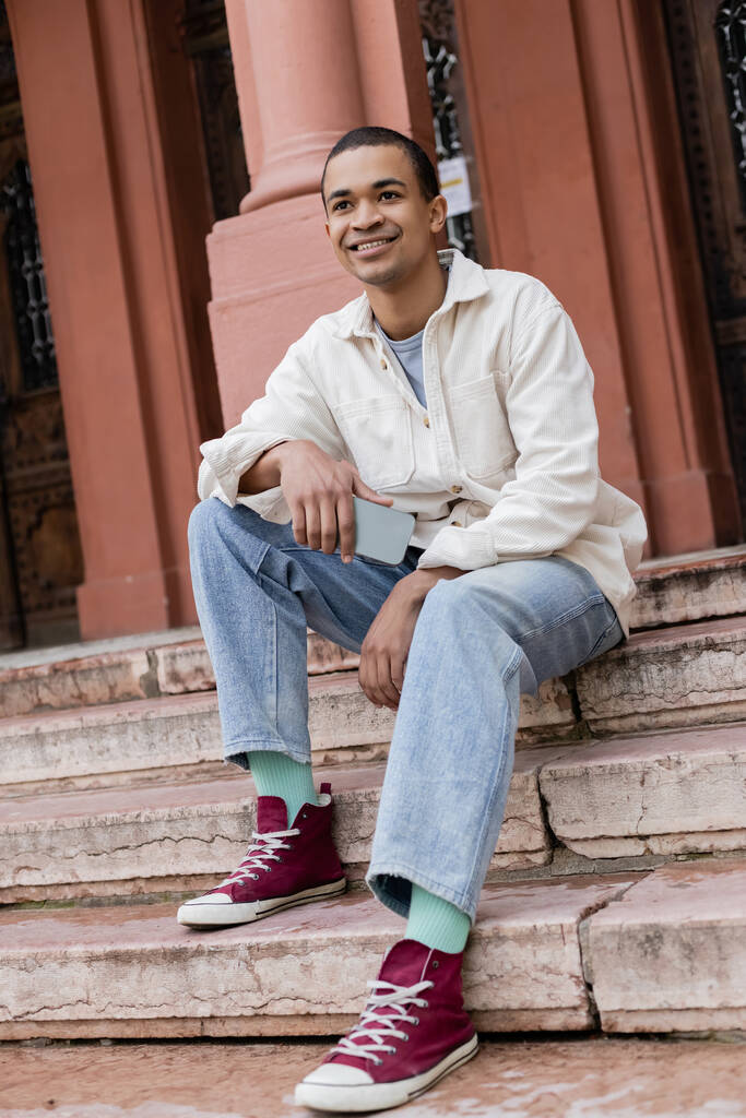 full length of smile African american man in shirt jacket holding smartphone ενώ κάθεται σε σκάλες στην πόλη  - Φωτογραφία, εικόνα