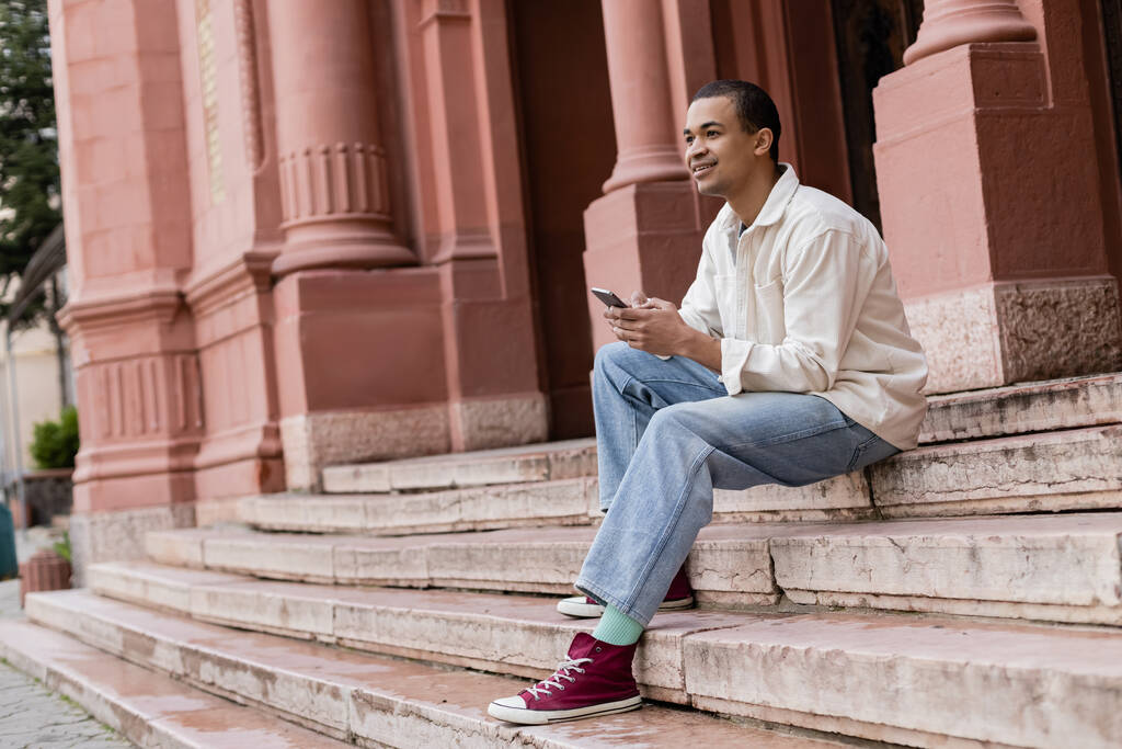 plná délka šťastný africký Američan muž v košili bunda drží mobilní telefon, zatímco sedí na schodech ve městě  - Fotografie, Obrázek