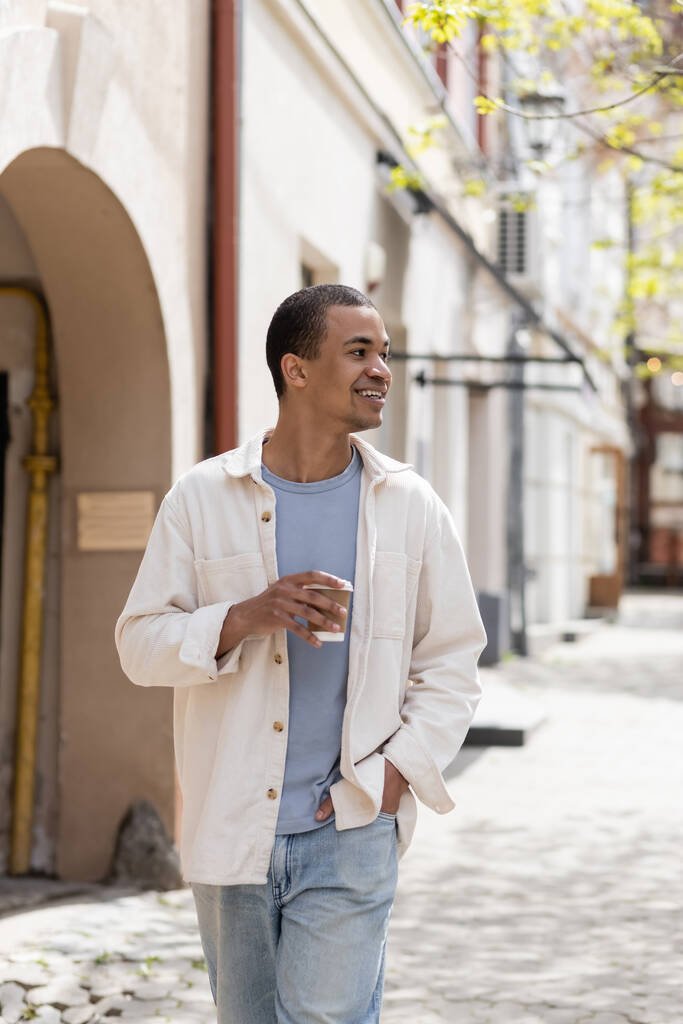 gelukkig Afrikaans amerikaanse man in shirt jas houden afhaaldrankje en lopen met de hand in zak in stedelijke stad  - Foto, afbeelding