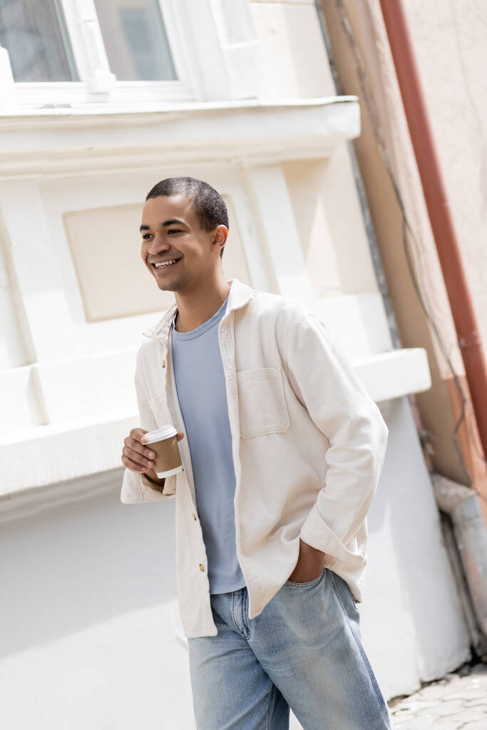 シャツに身を包んだアフリカ系アメリカ人の陽気な男がコーヒーを持って都市部を歩き  - 写真・画像