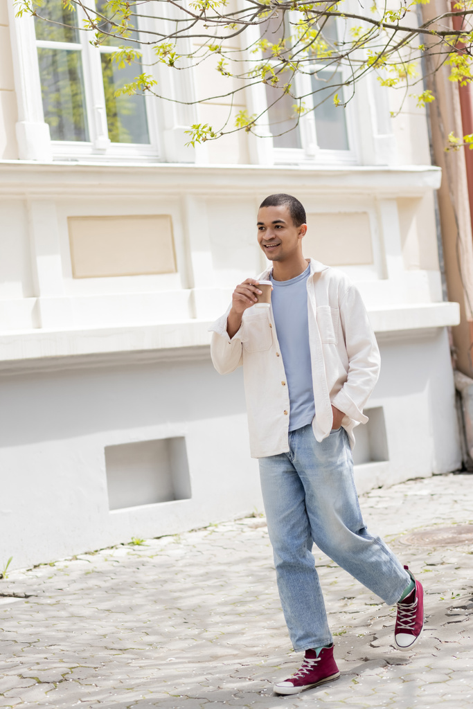 longitud completa de hombre afroamericano feliz en camisa chaqueta y pantalones vaqueros sosteniendo café para ir y caminar en la ciudad urbana - Foto, imagen