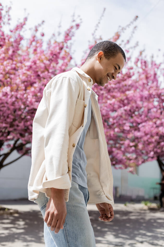 vue latérale de l'homme afro-américain heureux en veste chemise marchant près de cerisier rose - Photo, image