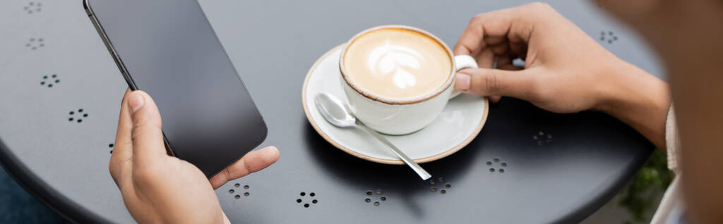 gedeeltelijke weergave van Afrikaanse Amerikaanse man sms 'en op smartphone in de buurt van cappuccino op tafel, banner - Foto, afbeelding