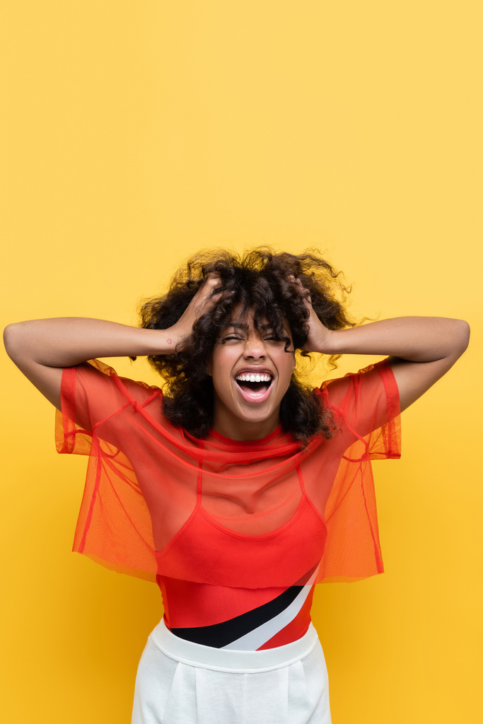 excité afro-américain femme avec les mains sur la tête riant avec les yeux fermés isolé sur jaune - Photo, image